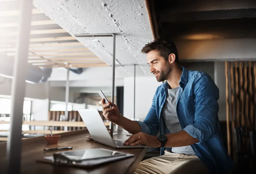 Small business owner smiling at his phone while working on a laptop in a café